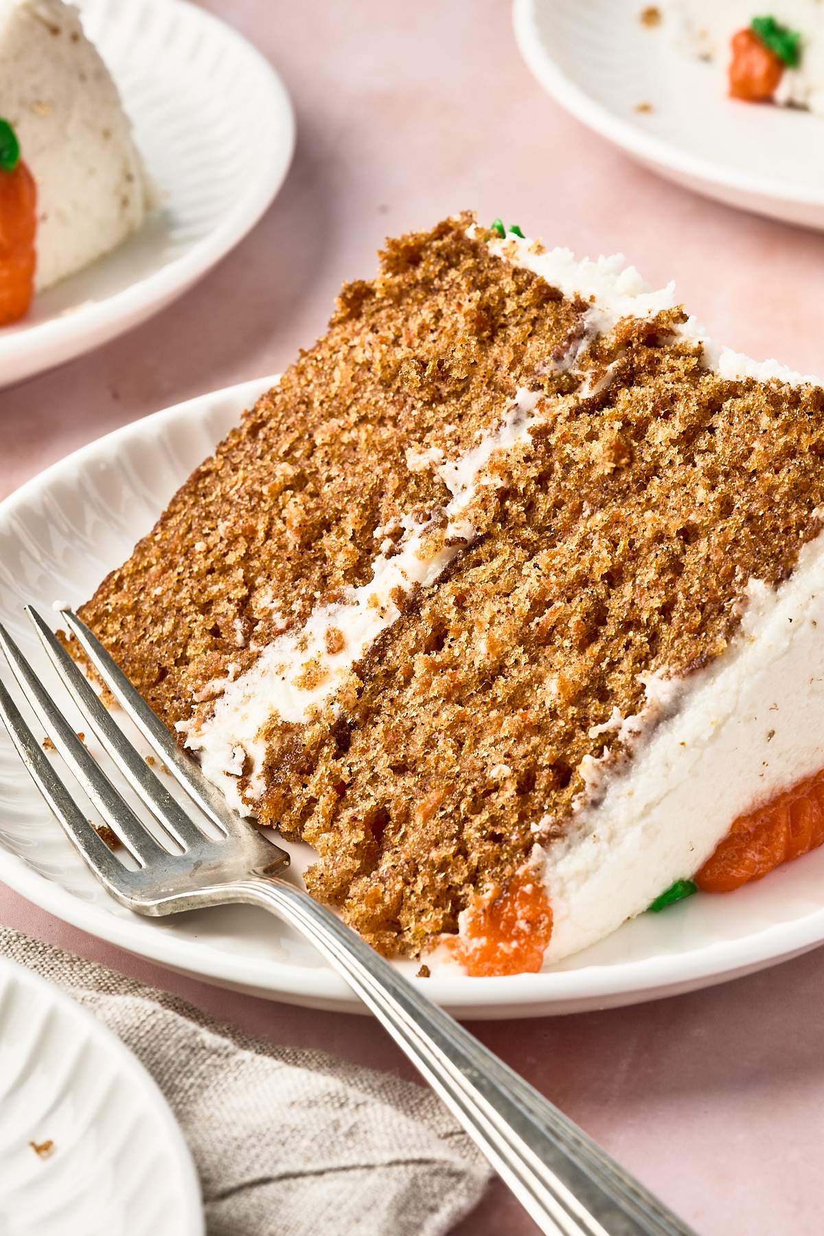 A close-up of a slice of carrot cake with white frosting on a white plate, accompanied by a fork. The background shows partial views of more cake slices on plates.