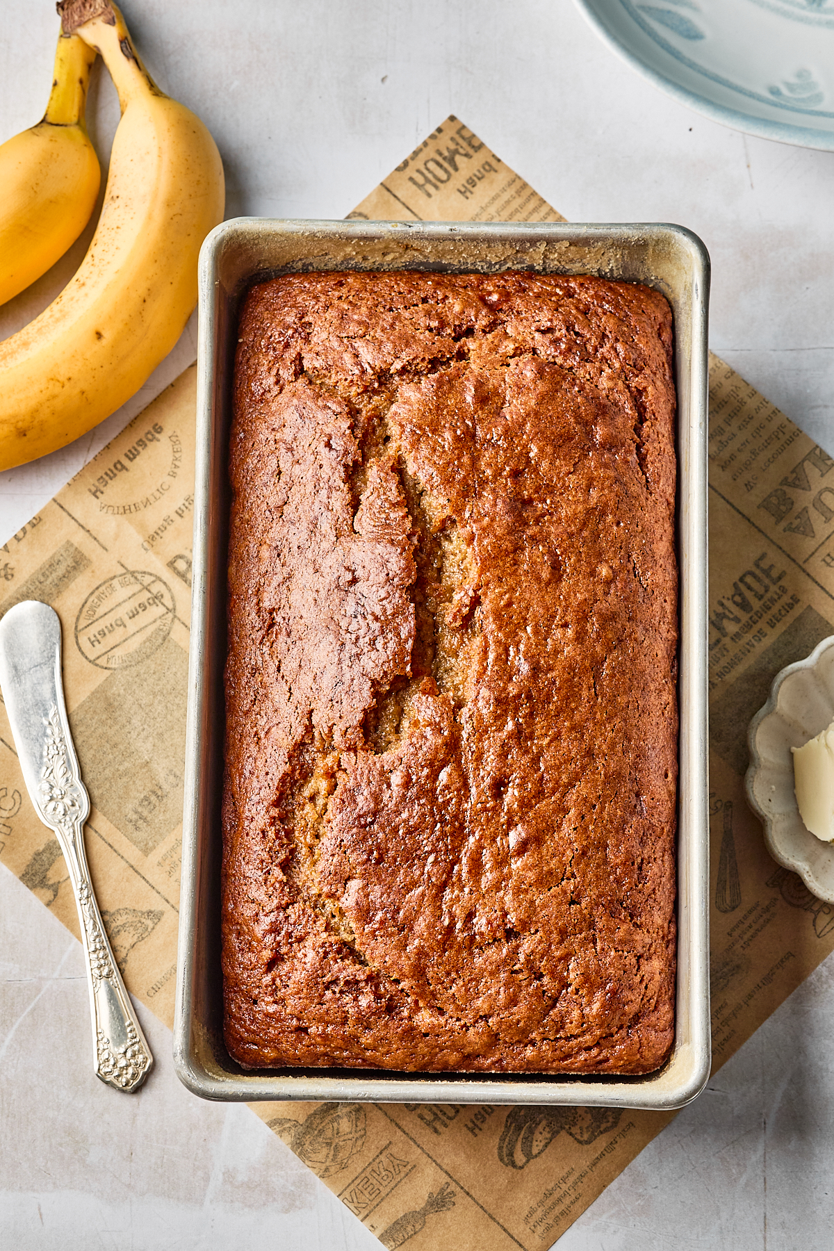 A loaf of banana bread in a rectangular baking pan sits on a parchment sheet, with two bananas and a vintage butter knife nearby, on a light-colored surface.
