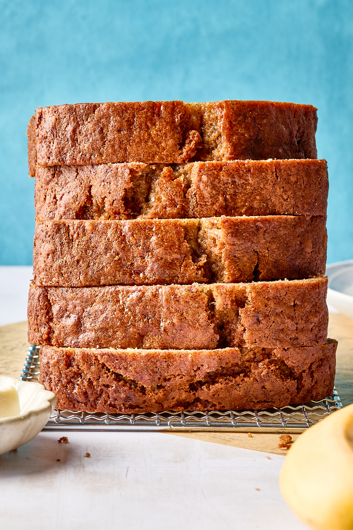 A stack of six thick slices of dairy free banana bread on a cooling rack, with a blue background and a pat of vegan butter on the side.
