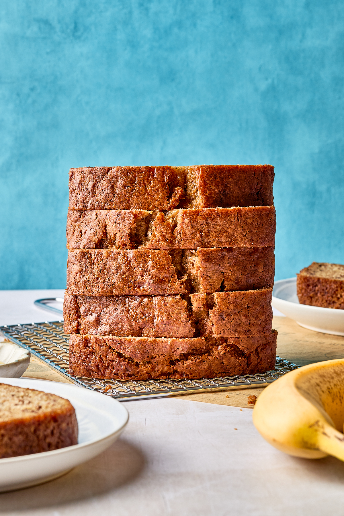 A stack of thick banana bread slices sits on a cooling rack, with plates of more banana bread and a ripe banana nearby, against a bright blue background.