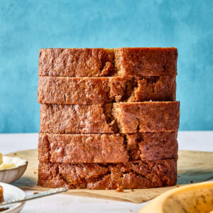 A stack of five thick slices of banana bread sits on a wooden surface, with a blue background and ripe bananas partially visible in the foreground.
