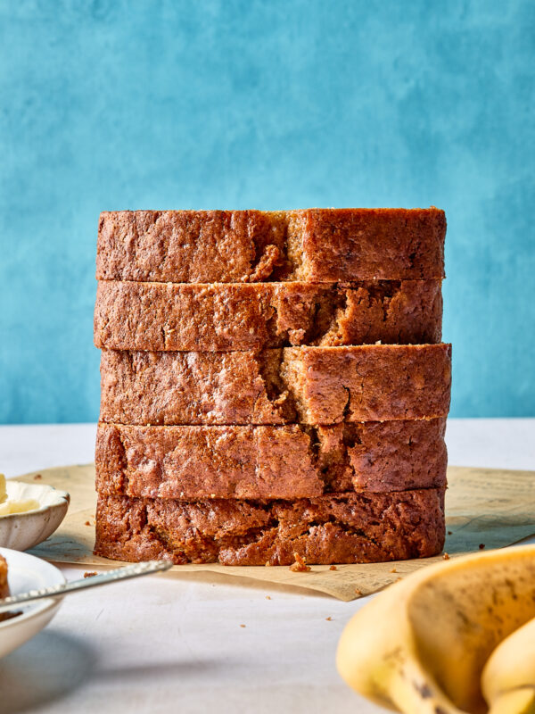 A stack of five thick slices of banana bread sits on a wooden surface, with a blue background and ripe bananas partially visible in the foreground.