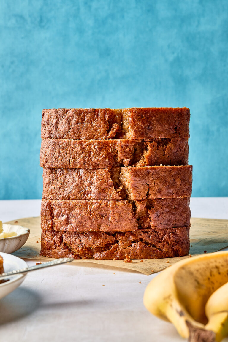 A stack of five thick slices of banana bread sits on a wooden surface, with a blue background and ripe bananas partially visible in the foreground.