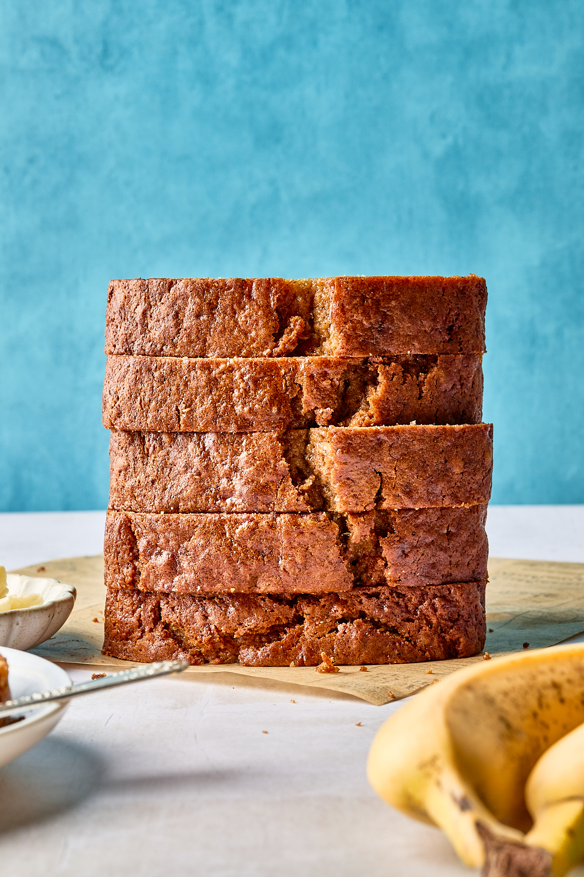 A stack of five thick slices of banana bread sits on a wooden surface, with a blue background and ripe bananas partially visible in the foreground.