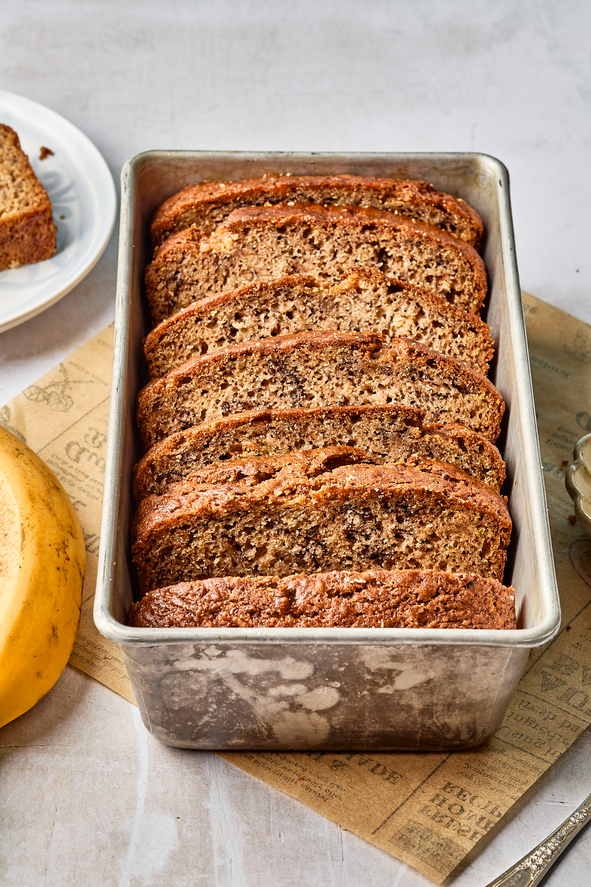 A loaf pan filled with neatly sliced dairy free banana bread sits on a brown sheet of paper, next to a ripe banana and a small dish. One slice is on a white plate in the background.