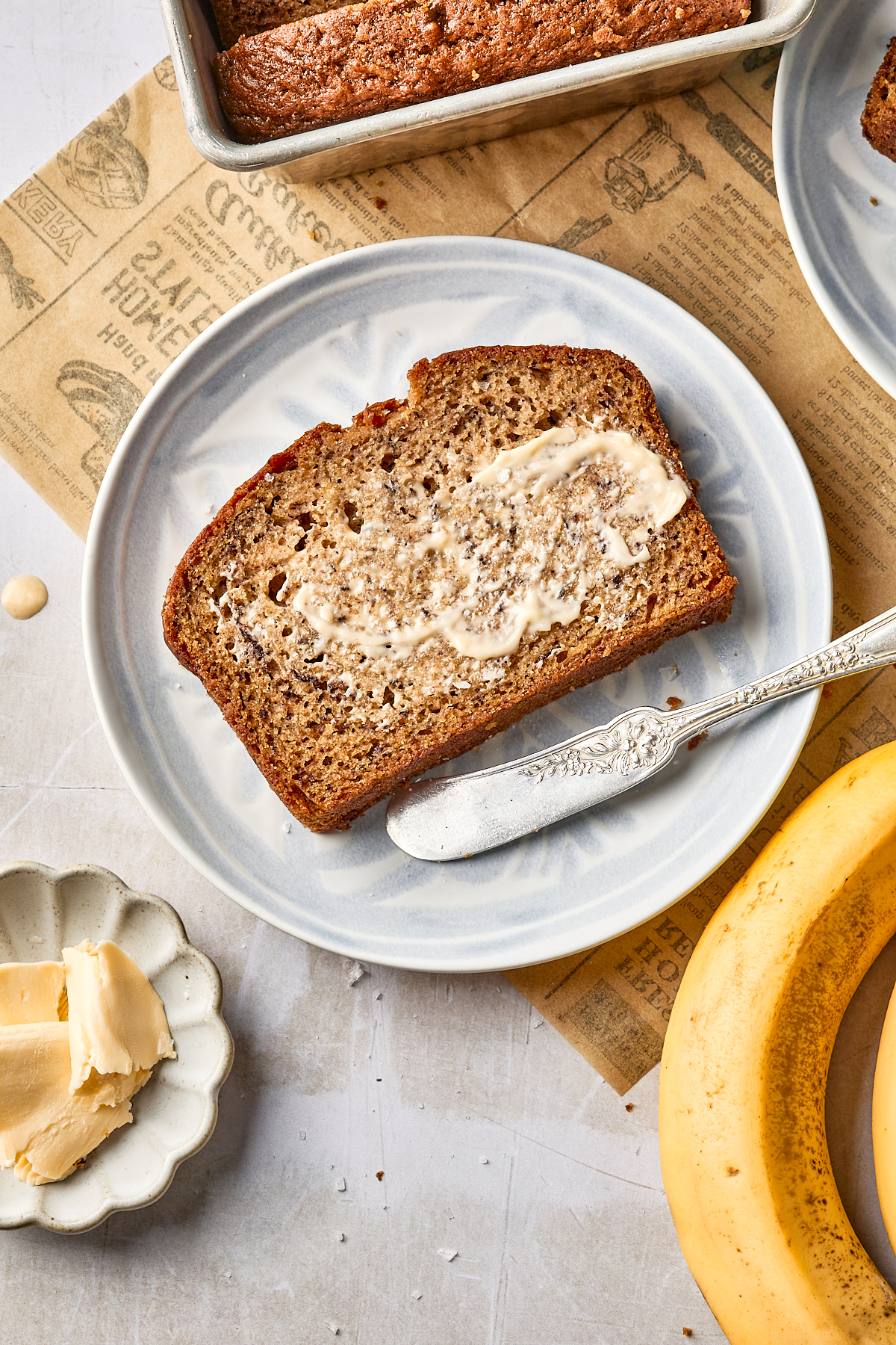 A slice of banana bread with melted dairy free butter is on a white plate next to a butter knife. Nearby are a pat of butter, a ripe banana, and part of a loaf still in its pan.