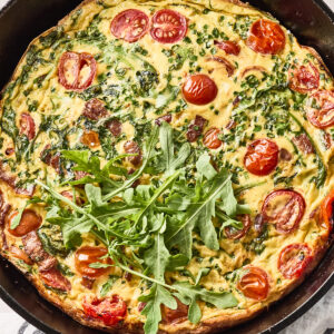 A baked frittata in a cast iron skillet, topped with fresh arugula, with visible cherry tomatoes, herbs, and leafy greens mixed into the egg. The skillet sits on a striped cloth, surrounded by fresh ingredients.