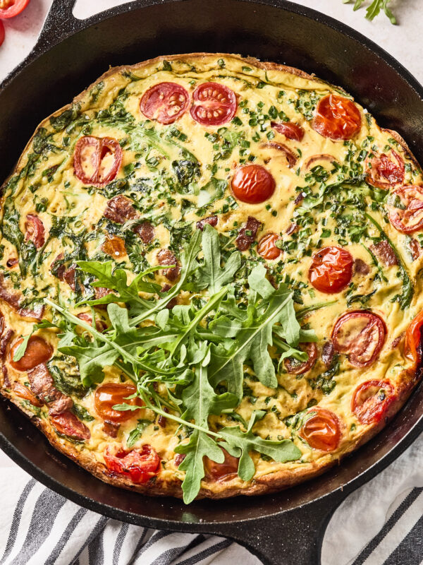 A baked frittata in a cast iron skillet, topped with fresh arugula, with visible cherry tomatoes, herbs, and leafy greens mixed into the egg. The skillet sits on a striped cloth, surrounded by fresh ingredients.