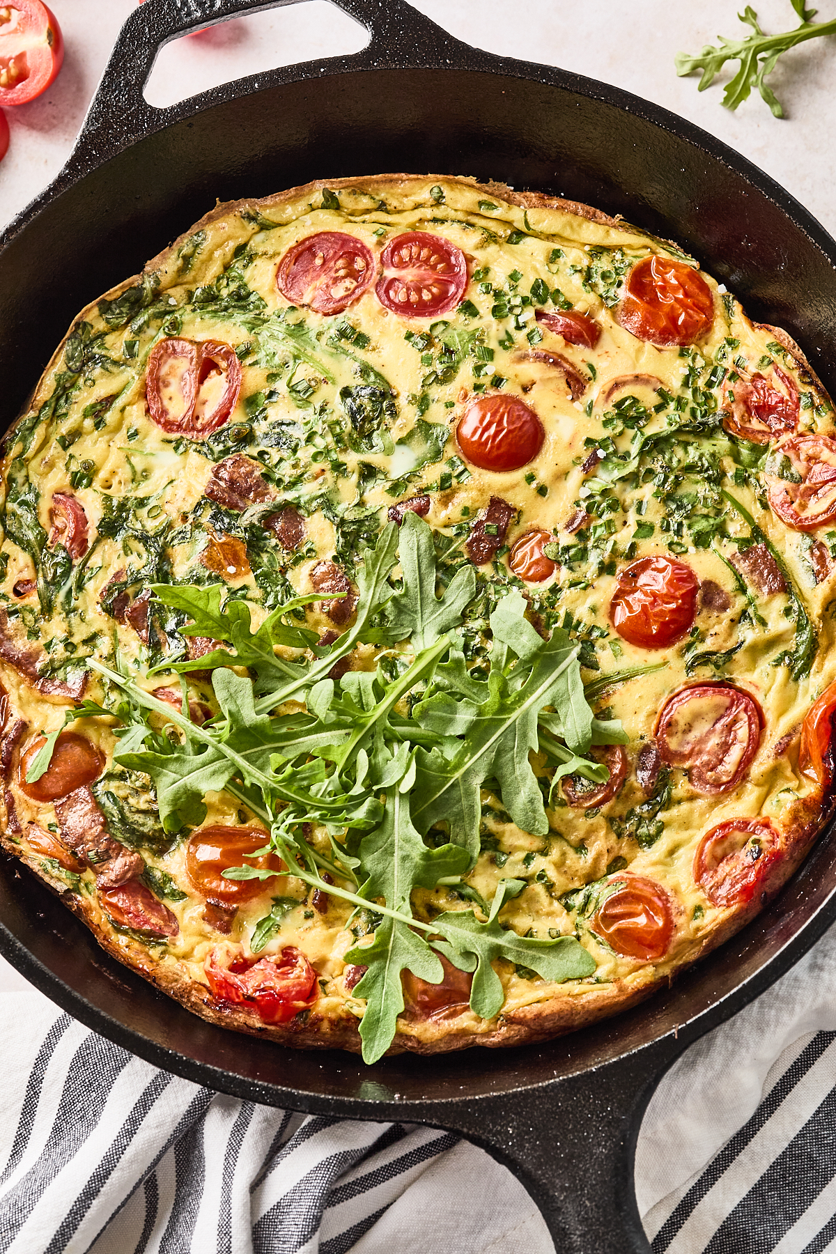 A baked frittata in a cast iron skillet, topped with fresh arugula, with visible cherry tomatoes, herbs, and leafy greens mixed into the egg. The skillet sits on a striped cloth, surrounded by fresh ingredients.