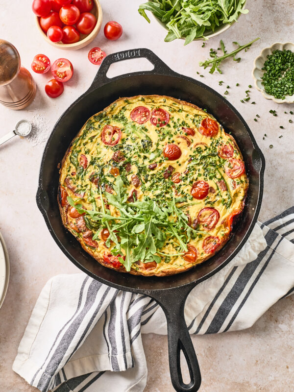 A baked frittata with cherry tomatoes and chopped herbs sits in a black cast iron skillet, garnished with fresh arugula. Surrounding the skillet are bowls of cherry tomatoes, arugula, chives, and a pepper grinder.
