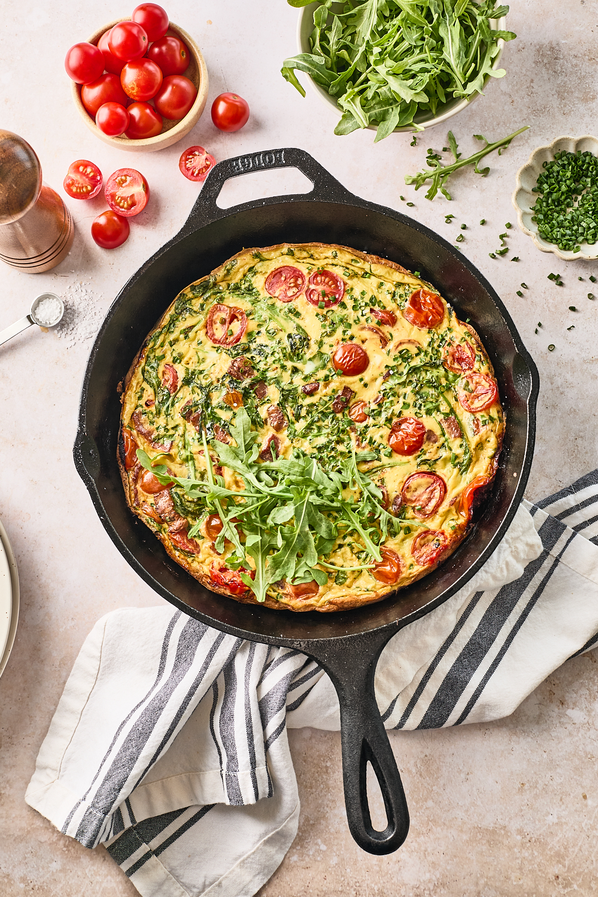 A baked frittata with cherry tomatoes and chopped herbs sits in a black cast iron skillet, garnished with fresh arugula. Surrounding the skillet are bowls of cherry tomatoes, arugula, chives, and a pepper grinder.