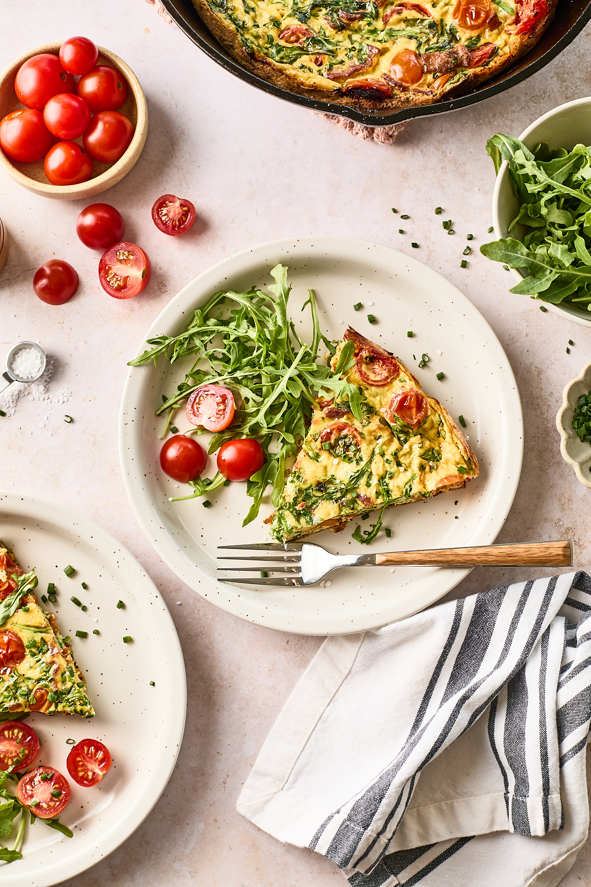 A plate with a slice of vegetable frittata, cherry tomatoes, and arugula. Nearby are more plates, a bowl of cherry tomatoes, a bowl of arugula, a skillet with frittata, and a striped napkin with a fork.