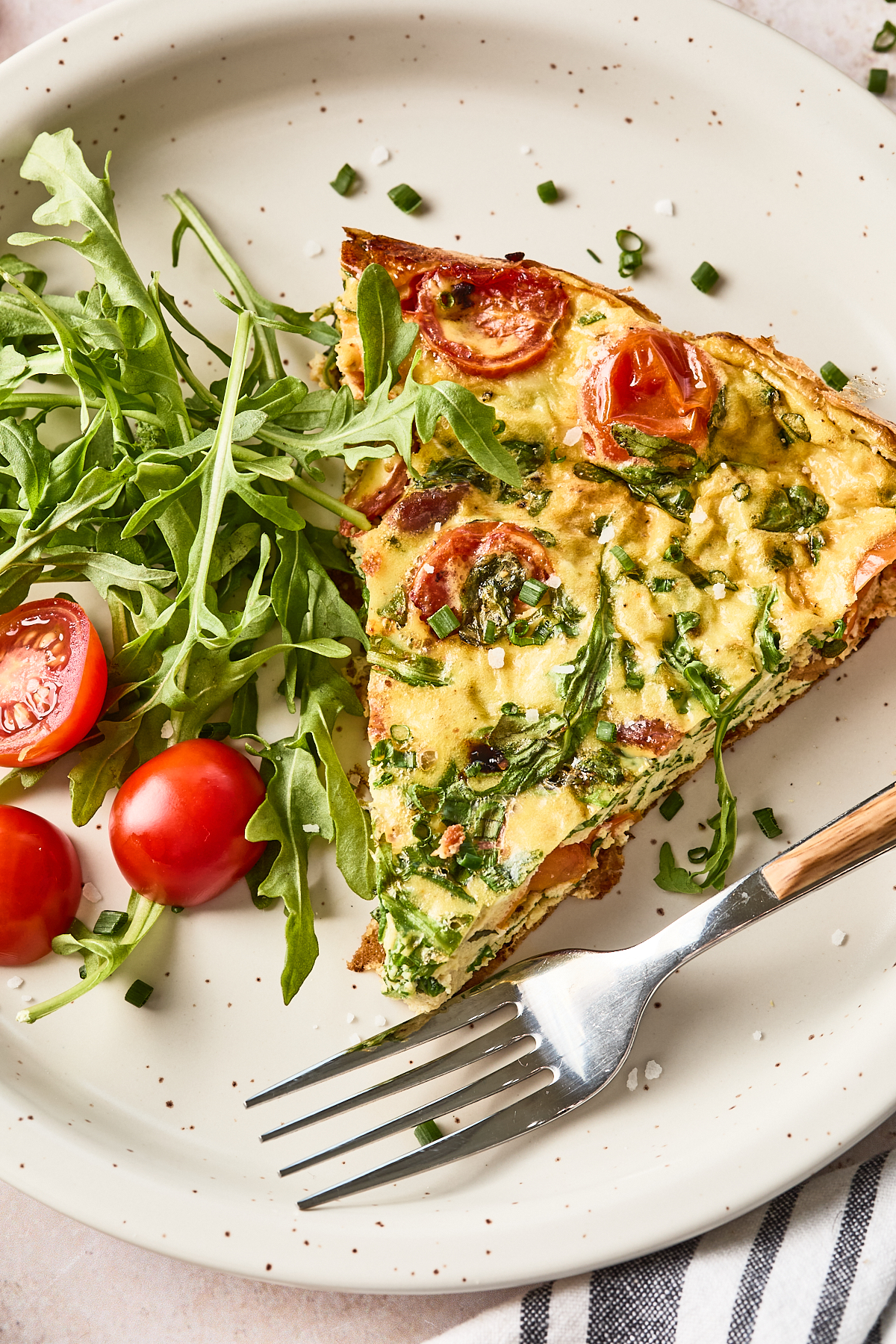 A slice of vegetable frittata with tomatoes and greens is served on a white plate, garnished with arugula and cherry tomatoes, with a fork placed beside the food.
