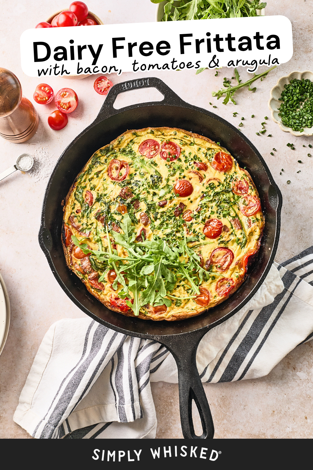 A cast iron skillet filled with a dairy-free frittata topped with arugula and cherry tomatoes sits on a striped towel, surrounded by fresh ingredients. Text reads: “Dairy Free Frittata with bacon, tomatoes & arugula.”.