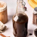 A glass bottle filled with dark liquid sits on a white surface, surrounded by coffee beans, cinnamon sticks, a jar with cinnamon sticks, a frothy iced coffee, and a bunch of bananas in the background.