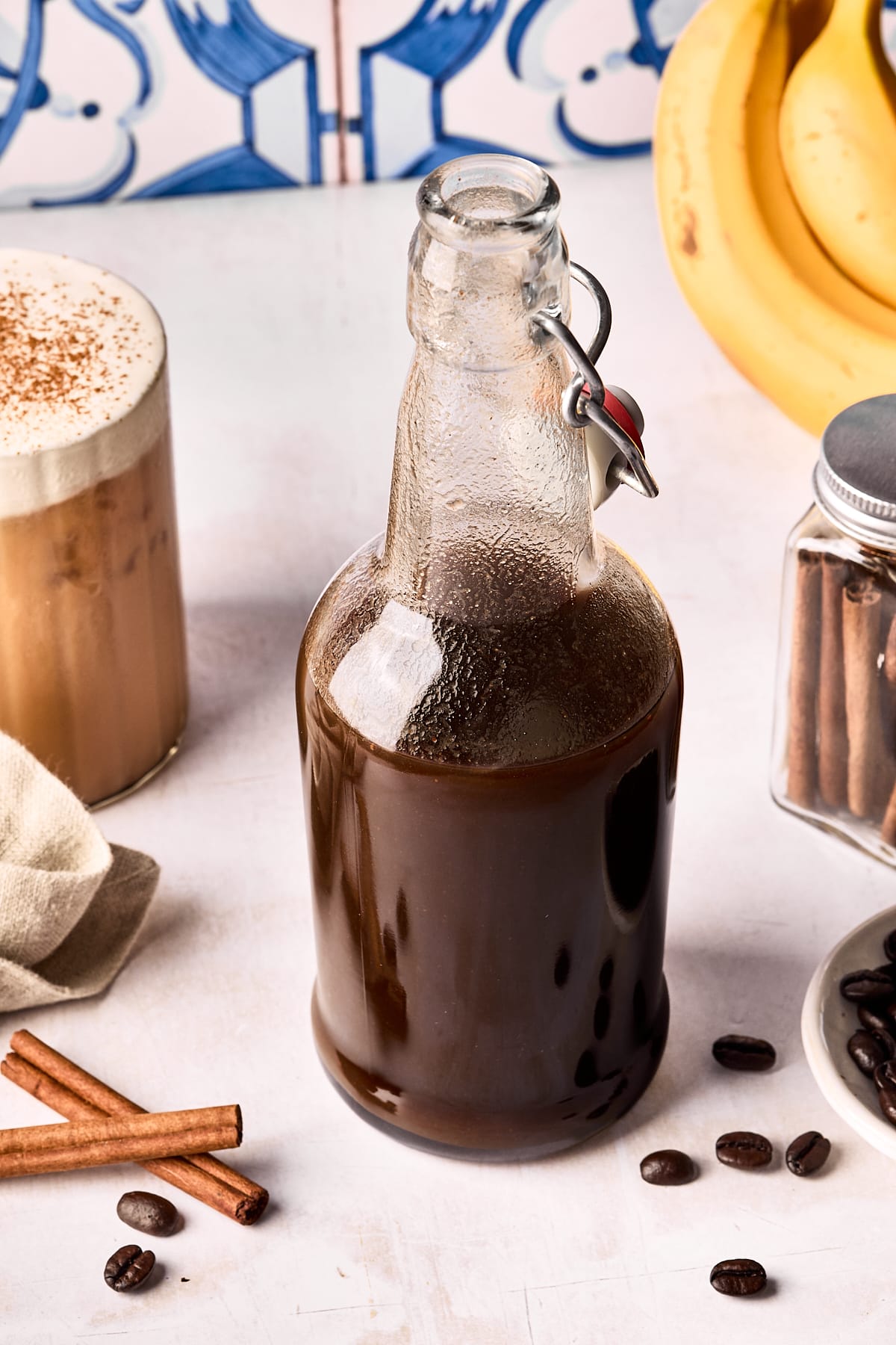 A glass bottle filled with dark liquid sits on a white surface, surrounded by coffee beans, cinnamon sticks, a jar with cinnamon sticks, a frothy iced coffee, and a bunch of bananas in the background.