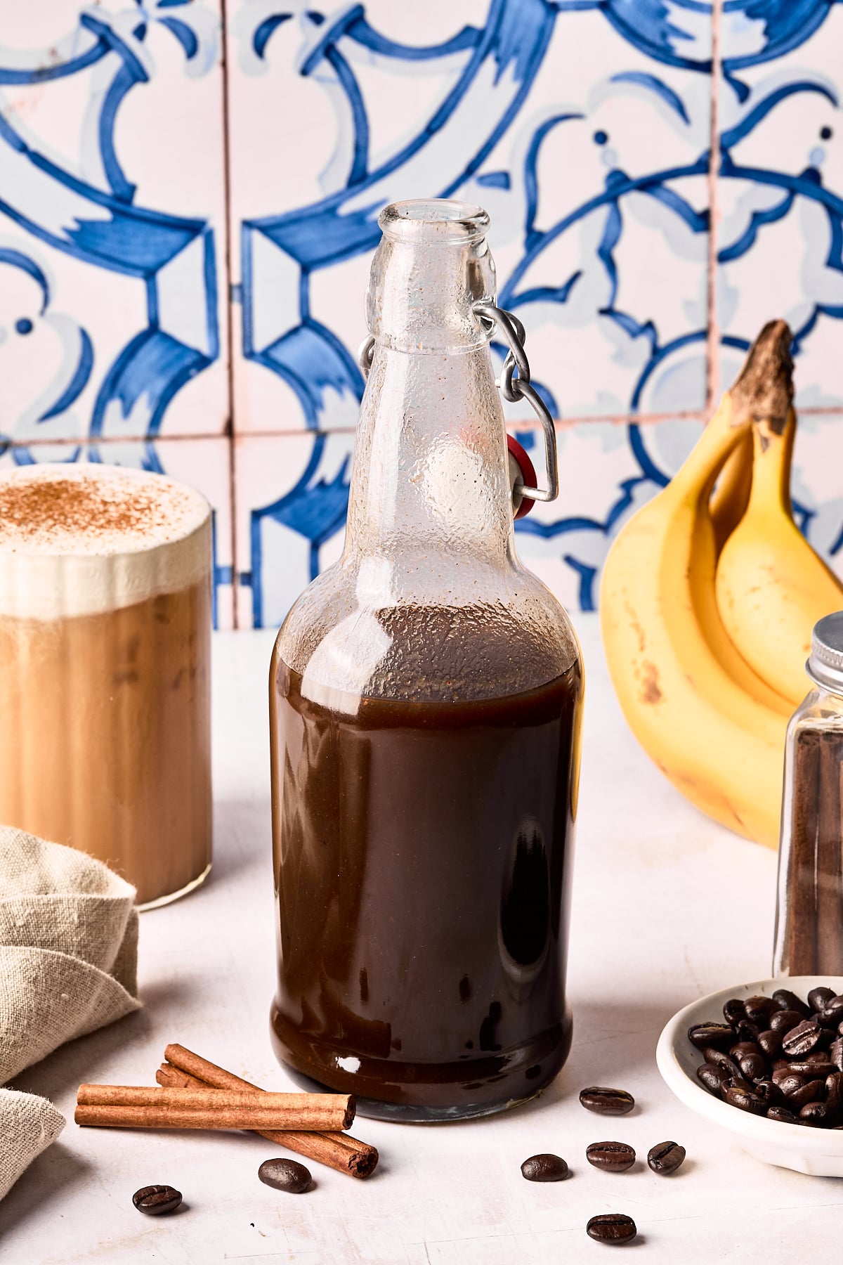 A glass bottle filled with dark liquid stands on a white surface next to bananas, a glass of iced coffee with foam, cinnamon sticks, coffee beans, and a jar. Blue and white patterned tiles are in the background.