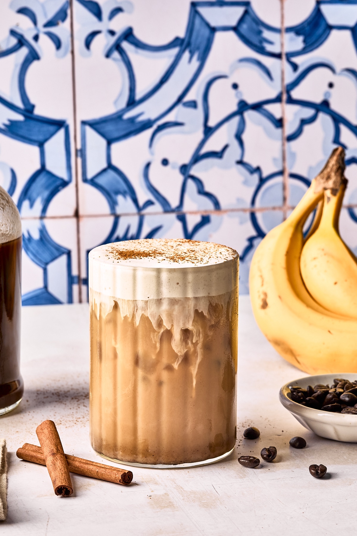 A creamy iced coffee drink topped with foam and cinnamon sits on a white surface. Nearby are a bunch of bananas, a bowl of coffee beans, cinnamon sticks, and a bottle, with blue patterned tiles in the background.