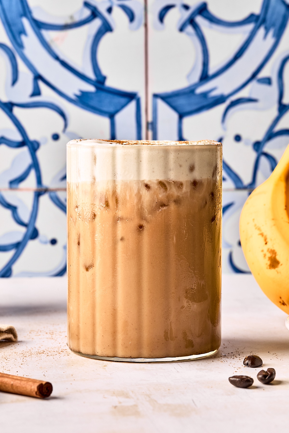 A glass of iced coffee with a thick layer of foam on top sits on a light surface. A banana, coffee beans, and a cinnamon stick are nearby, with a blue and white patterned tile in the background.