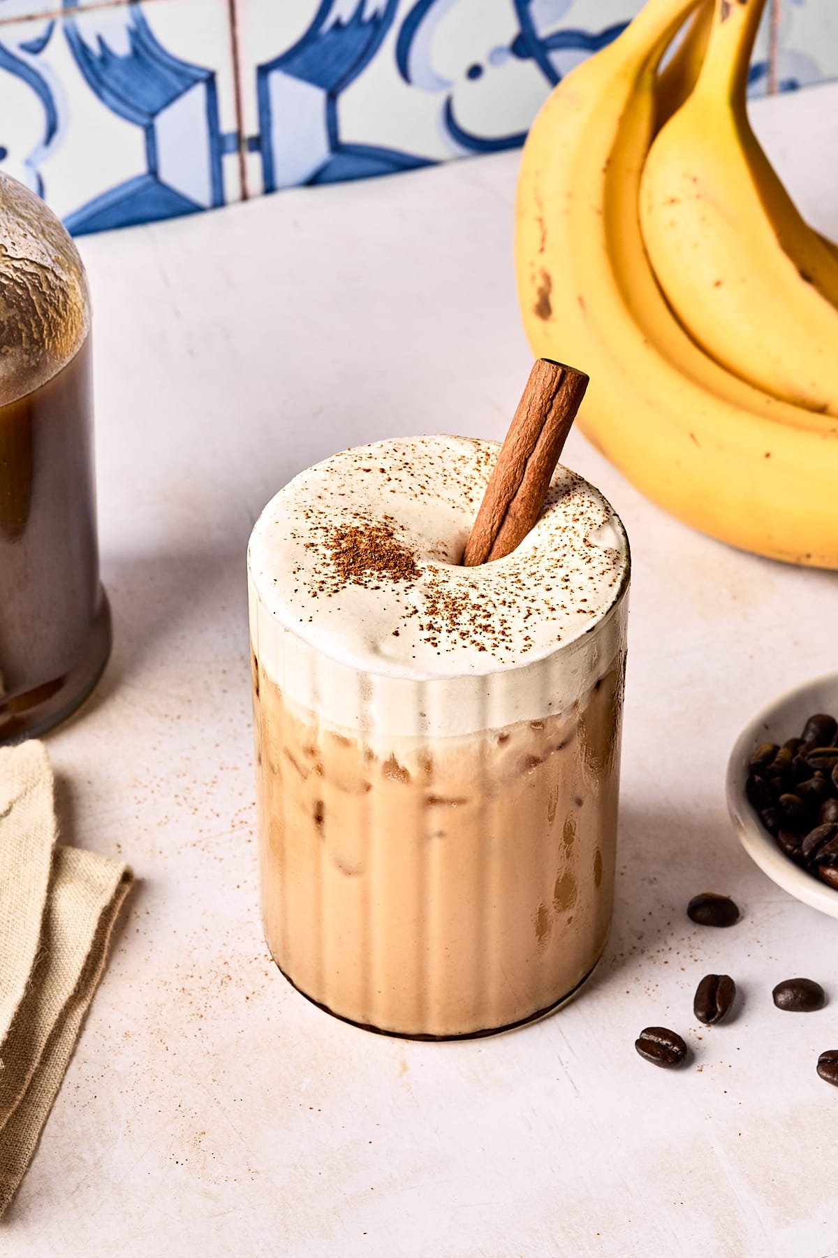 A glass of iced coffee with frothy cream on top, garnished with a cinnamon stick and a sprinkle of cinnamon. Bananas, coffee beans, and a bottle of coffee are nearby on a light surface. Blue patterned tiles are in the background.