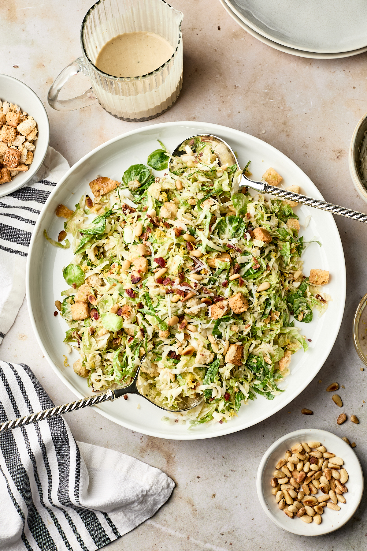 A plate of shredded Brussels sprout salad with croutons, nuts, and bacon bits, served with two serving spoons. Surrounding the plate are a bowl of pine nuts, a cup of dressing, and stacked empty plates on a light surface.
