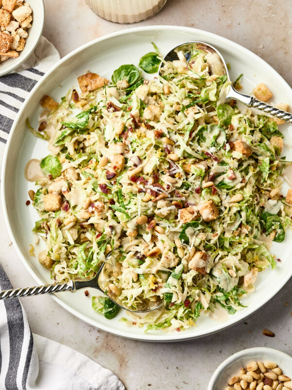A large white platter holds a fresh salad with shredded greens, croutons, grated cheese, and seeds, served with two silver serving spoons. A striped napkin and small bowl of seeds are nearby.