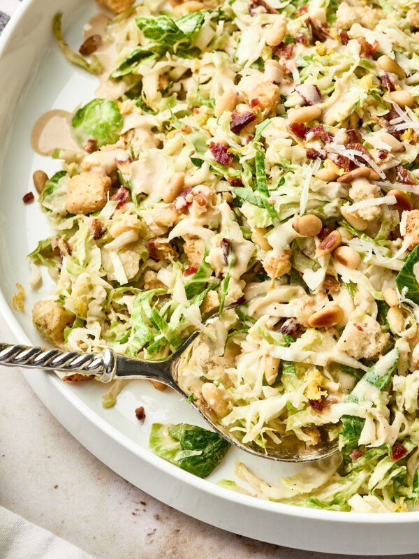 A close-up of a Caesar salad on a white plate, topped with shredded lettuce, pine nuts, croutons, bacon bits, grated cheese, and drizzled with creamy dressing. A serving spoon rests in the salad.