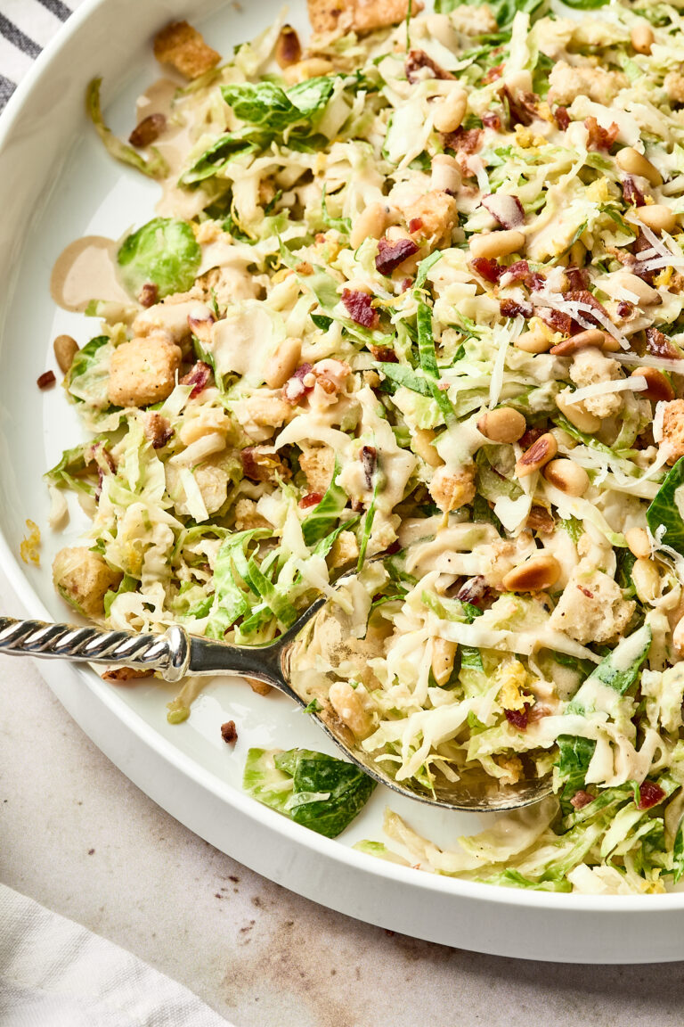 A close-up of a Caesar salad on a white plate, topped with shredded lettuce, pine nuts, croutons, bacon bits, grated cheese, and drizzled with creamy dressing. A serving spoon rests in the salad.