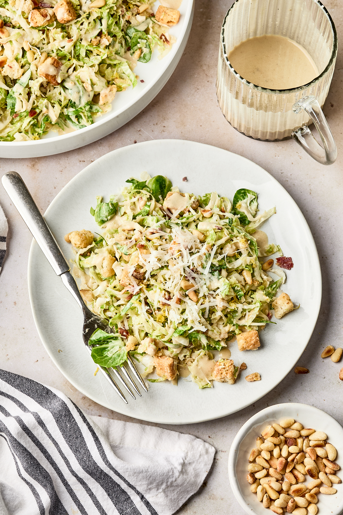 A plate of shredded Brussels sprouts salad with croutons, grated cheese, and creamy dressing sits beside a fork. A bowl of pine nuts, a glass cup of dressing, and a striped napkin are nearby.