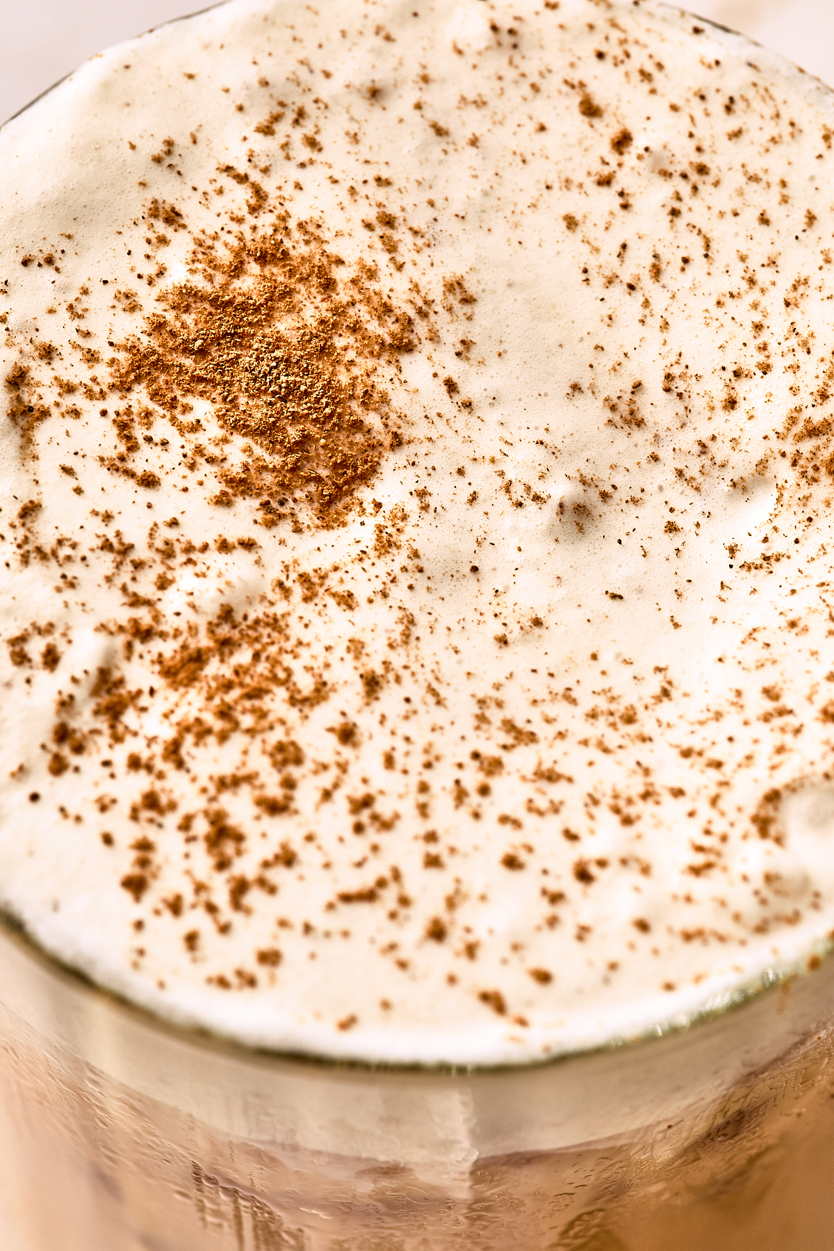 A close-up of a glass filled with frothy, creamy foam on top, sprinkled with ground cinnamon. The drink appears thick and rich, with the cinnamon scattered unevenly across the surface.