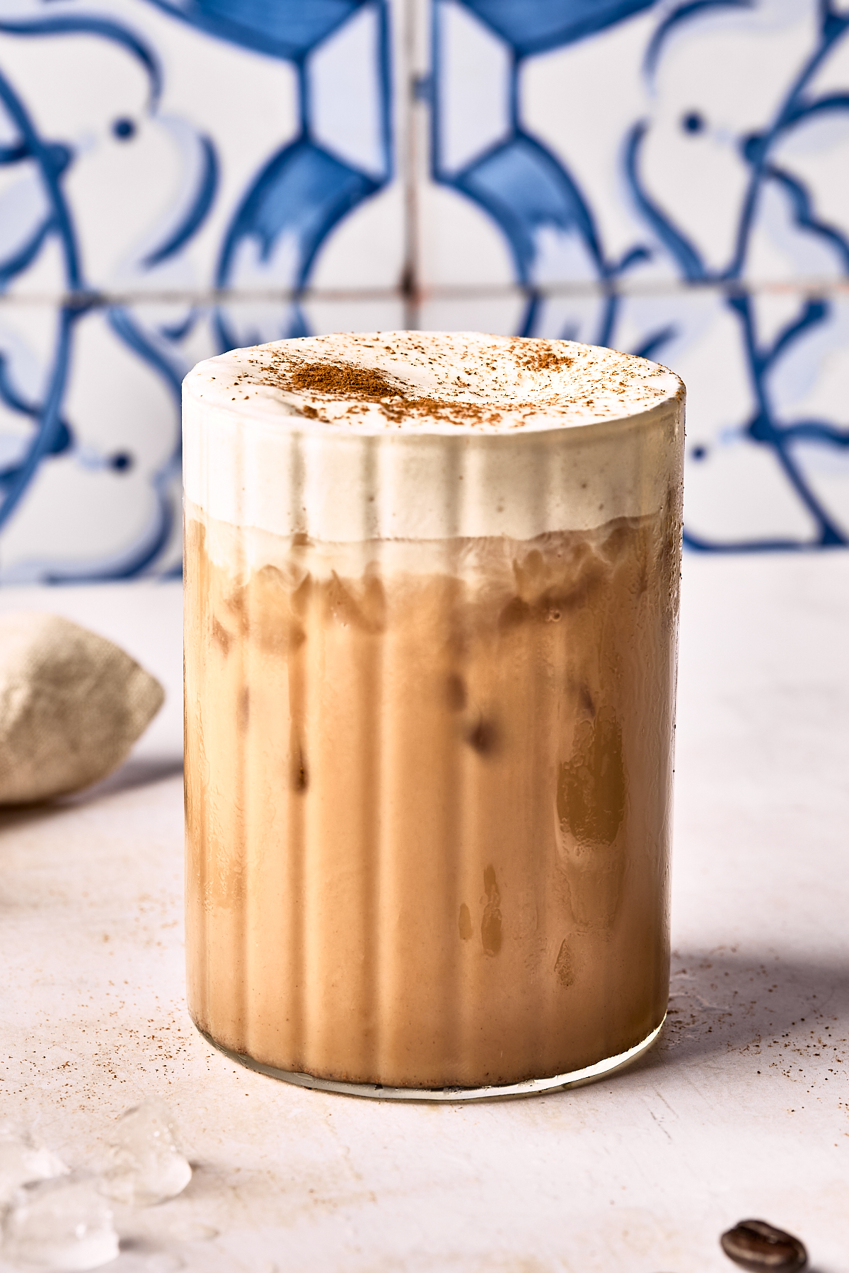 A glass of iced coffee topped with a thick layer of creamy foam and a sprinkle of cinnamon, set on a light surface with a blue and white tiled background.