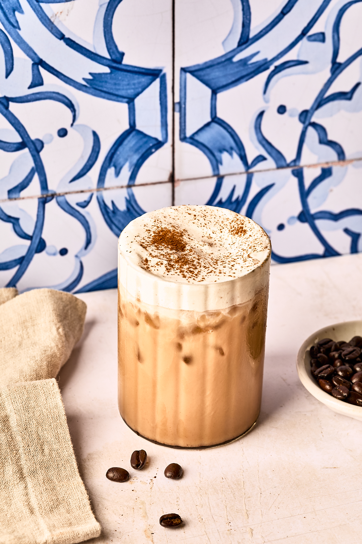 Iced coffee topped with frothy cream and a sprinkle of cinnamon, placed on a white surface with a beige napkin, coffee beans, and a small dish, against a blue patterned tile background.