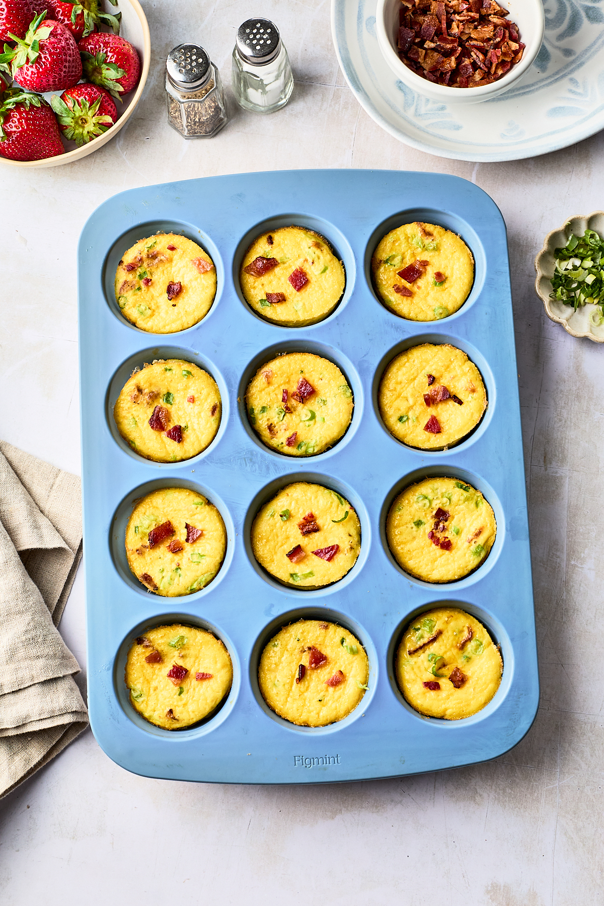 A blue muffin tin holds twelve baked egg bites with bits of bacon and green onions. Nearby are bowls of chopped bacon, sliced green onions, strawberries, salt and pepper shakers, and a folded beige napkin.