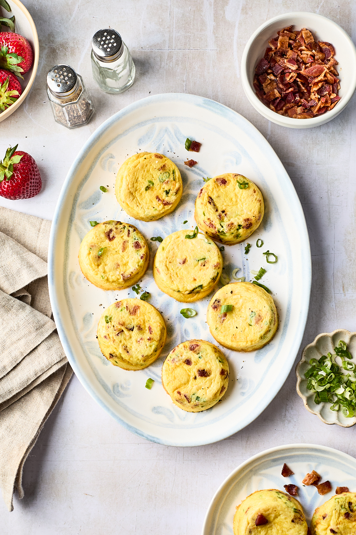 A white oval plate holds eight mini egg bites garnished with chopped green onions. Nearby are bowls of chopped bacon and more green onions, strawberries, salt and pepper shakers, and a beige napkin.