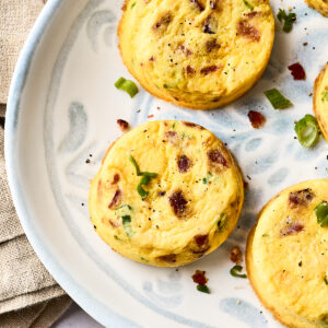 Three baked egg muffins with bits of bacon and green onion are arranged on a decorative white plate. A small dish with sliced green onions and a beige napkin are nearby.
