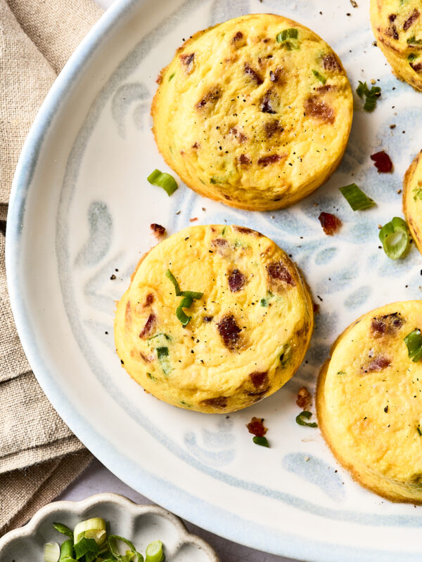 Three baked egg muffins with bits of bacon and green onion are arranged on a decorative white plate. A small dish with sliced green onions and a beige napkin are nearby.