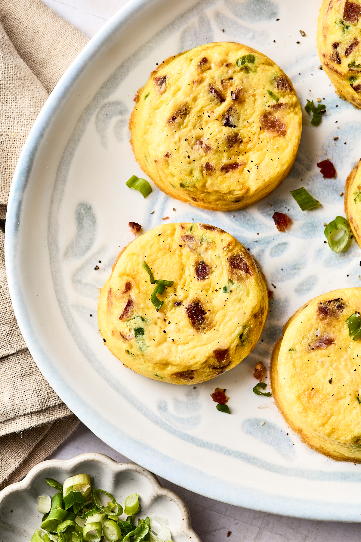 Three baked egg muffins with bits of bacon and green onion are arranged on a decorative white plate. A small dish with sliced green onions and a beige napkin are nearby.