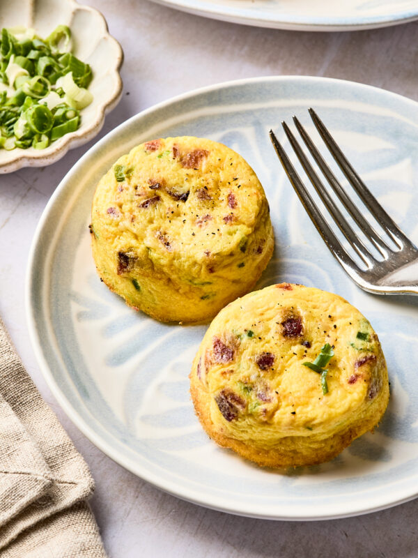 Two mini egg muffins with green herbs and pieces of bacon are served on a light blue plate next to a fork. A small dish with chopped green onions and a beige napkin are nearby.