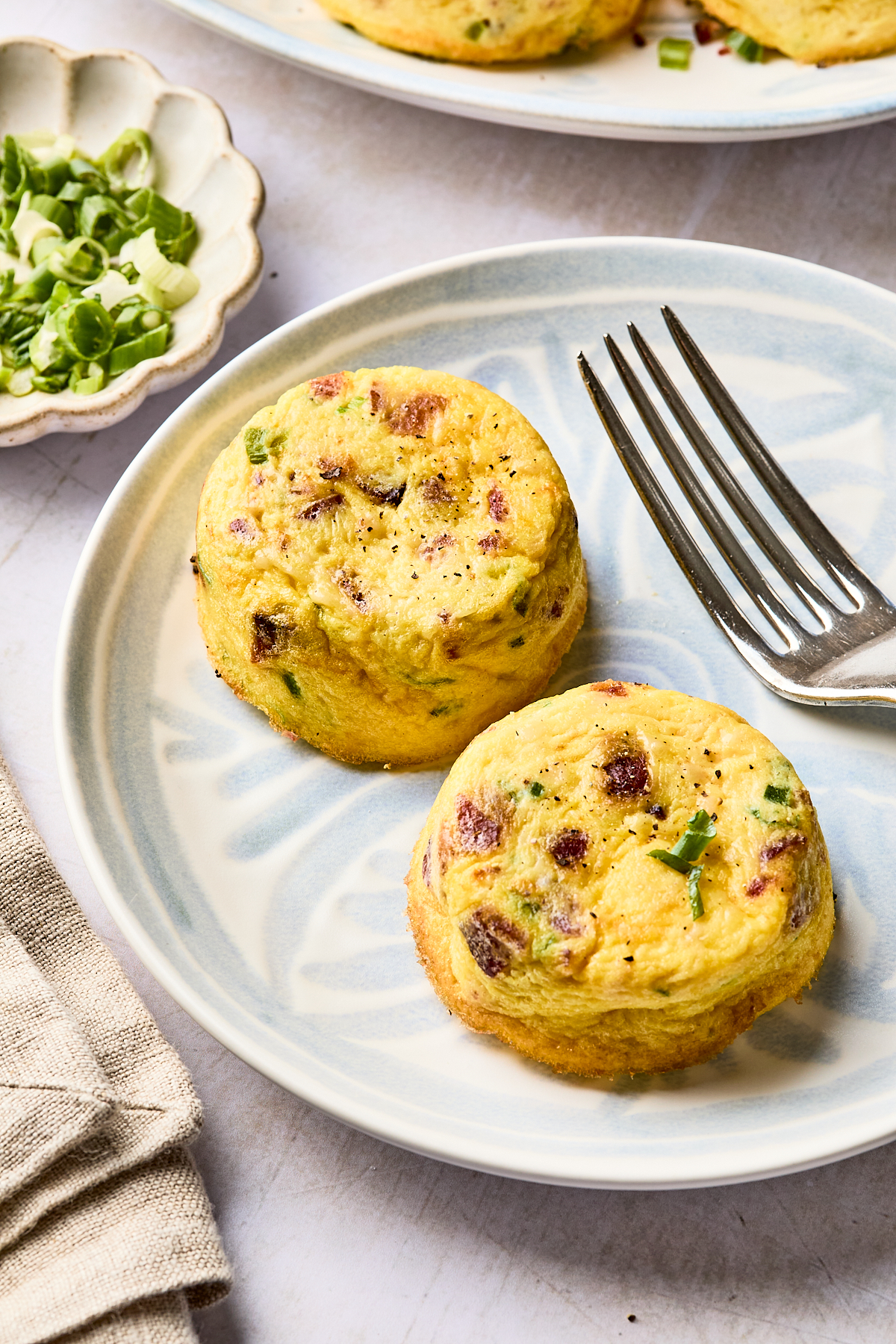 Two mini egg muffins with green herbs and pieces of bacon are served on a light blue plate next to a fork. A small dish with chopped green onions and a beige napkin are nearby.