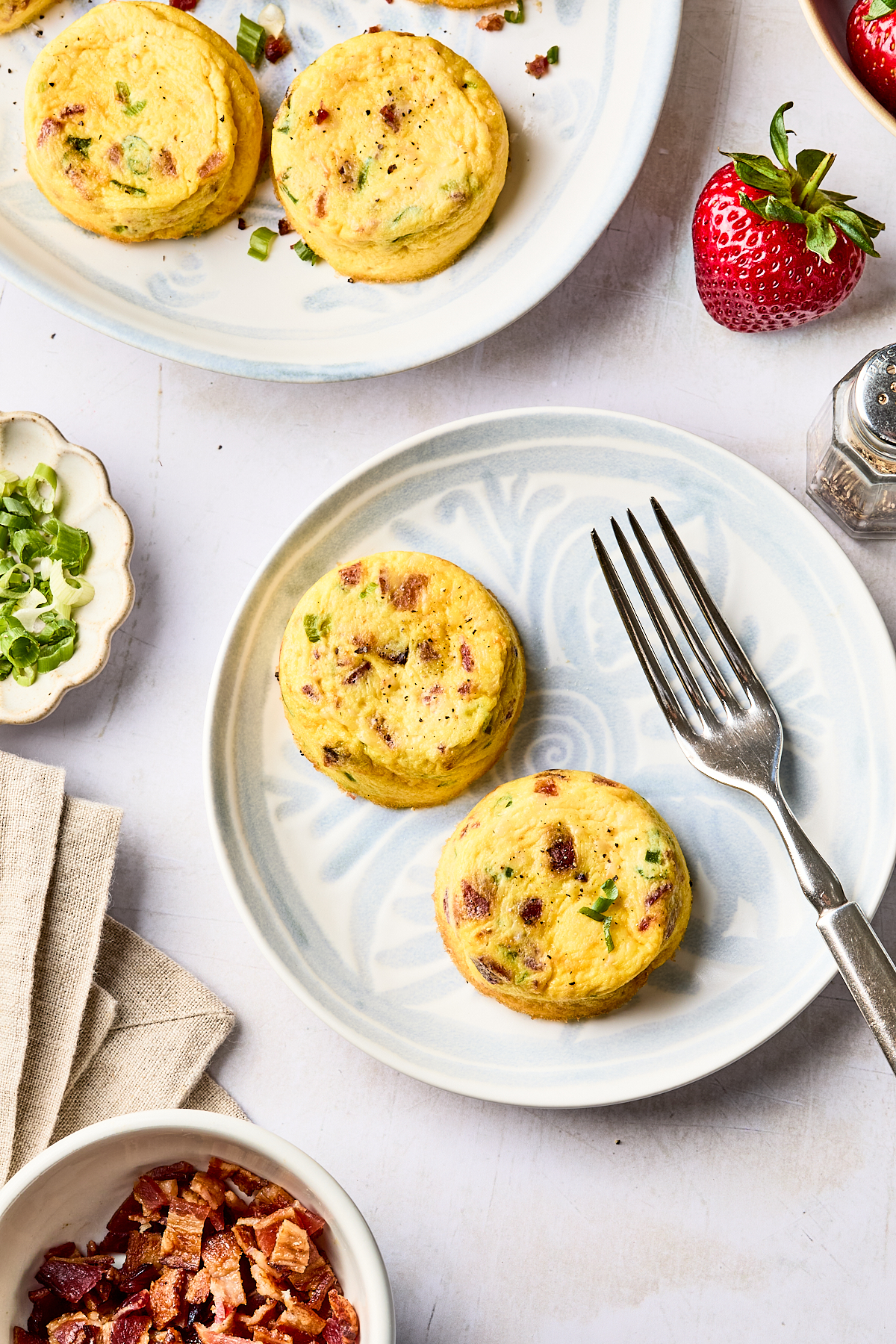 Two round egg muffins with herbs and bacon bits on a plate with a fork, surrounded by a strawberry, sliced green onions, bacon pieces, and another plate with more muffins.
