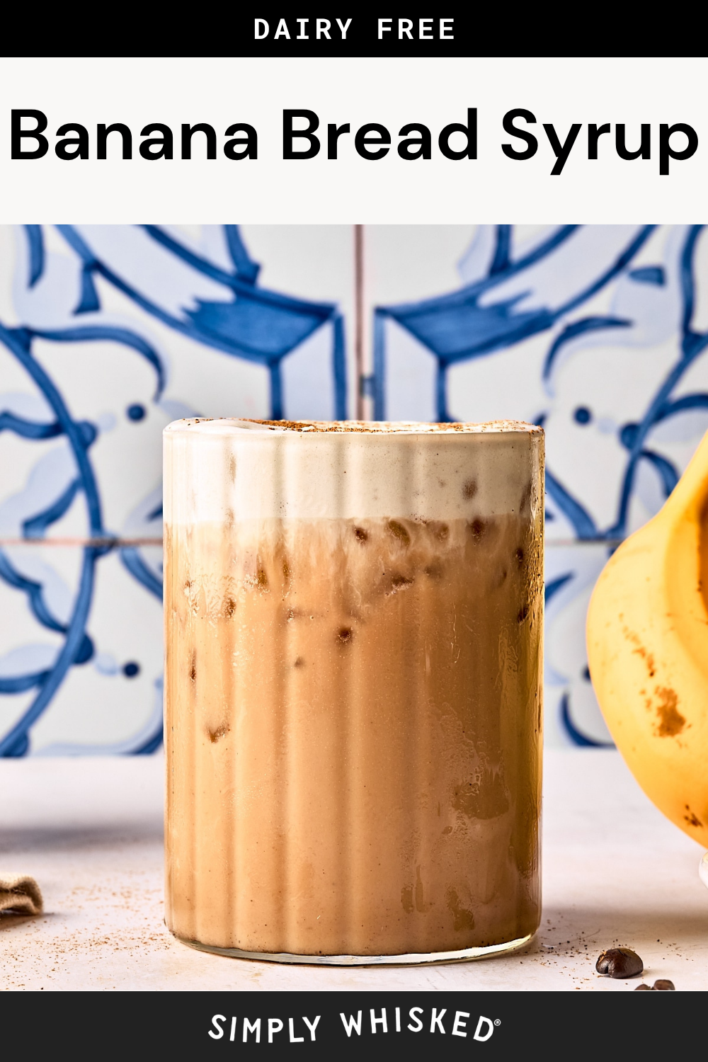 A glass of iced banana bread syrup drink topped with foam, set against a blue and white tile background. A banana and a pinch of cinnamon are visible beside the glass, highlighting the rich flavors of the signature banana bread syrup. Text reads Dairy Free Banana Bread Syrup.
