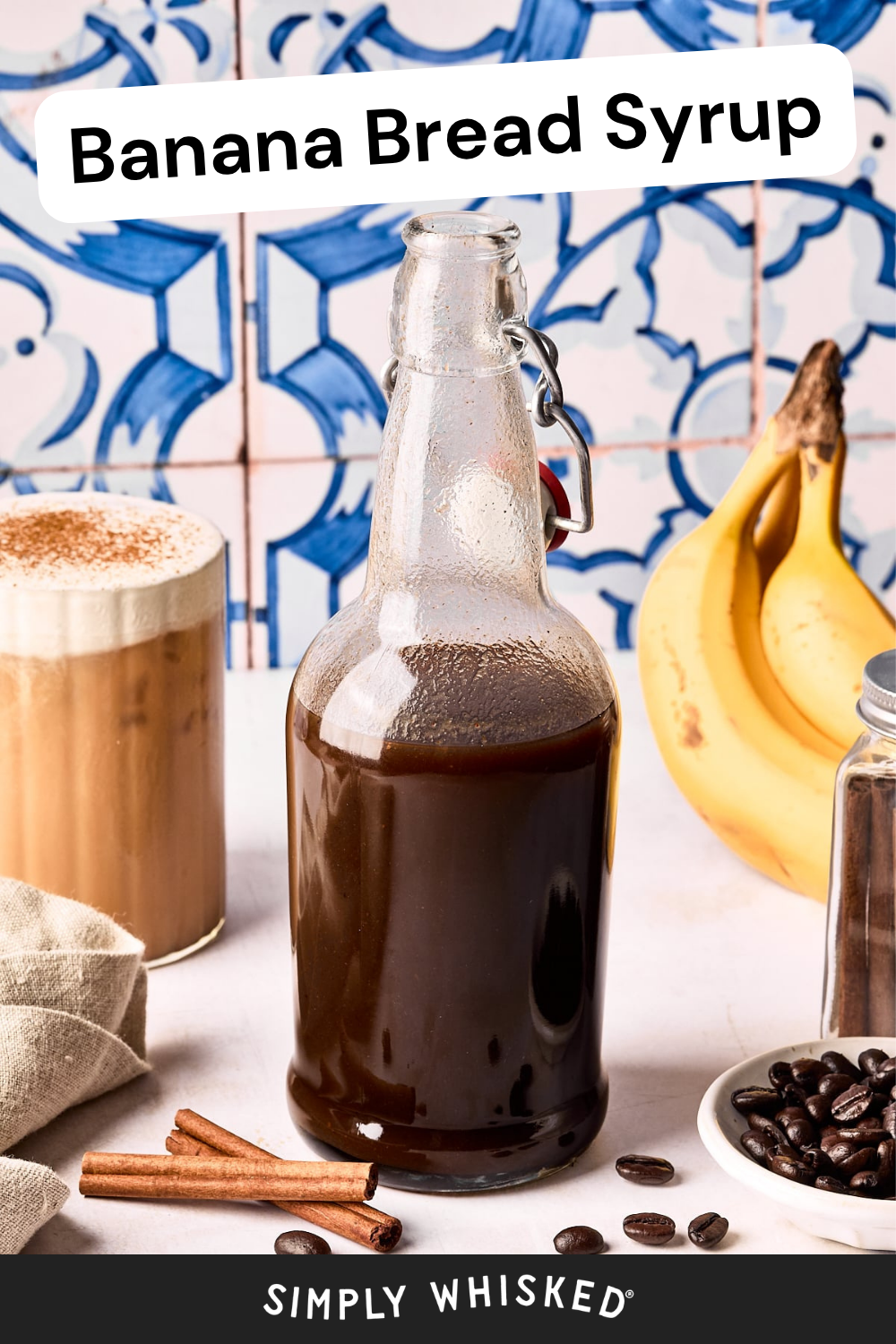 A glass bottle of banana bread syrup sits on a table with bananas, a coffee drink, coffee beans, cinnamon sticks, and a towel. A decorative blue and white tile wall is in the background. Text reads Banana Bread Syrup.