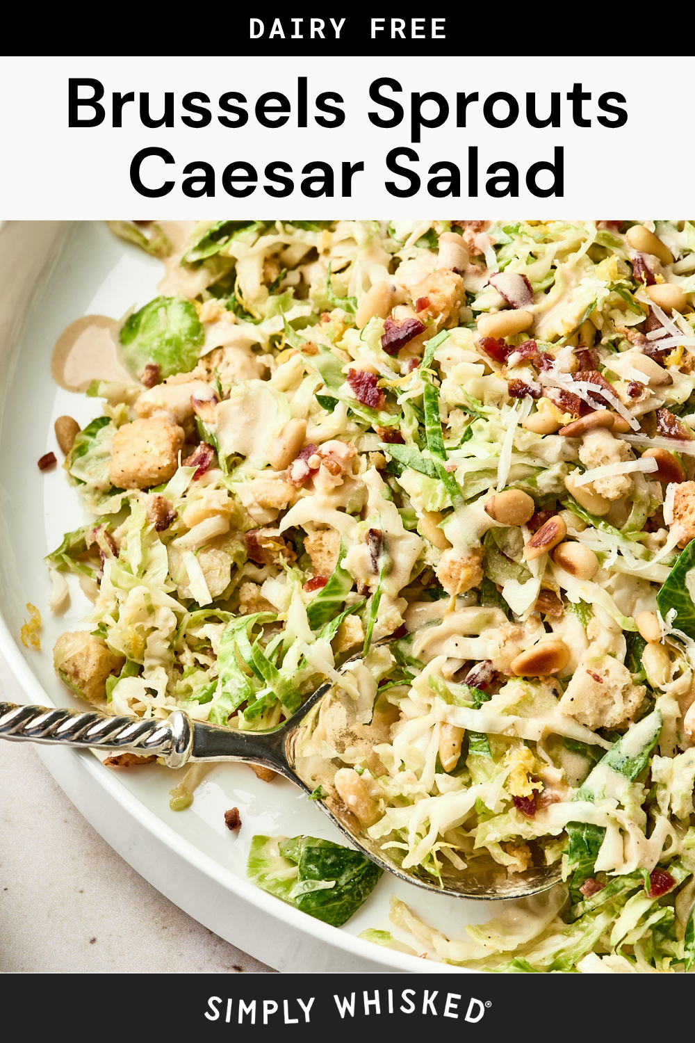 A close-up of a dairy-free brussels sprouts caesar salad with creamy dressing, croutons, and pine nuts on a white plate. A serving spoon rests in the salad. Text reads Brussels Sprouts Caesar Salad and Simply Whisked.