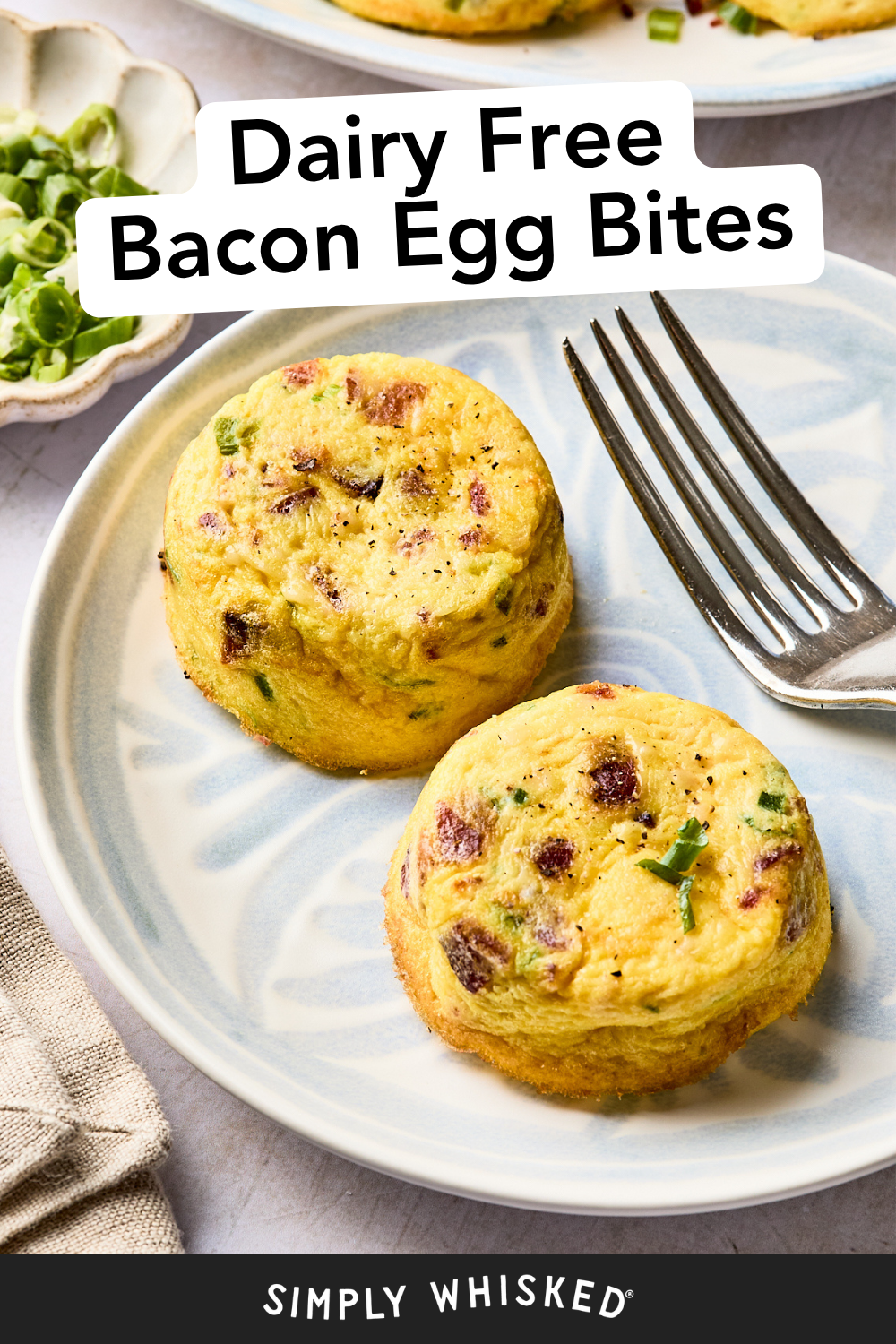 Two round dairy-free bacon egg bites are served on a light blue patterned plate with a fork. The bites appear fluffy with visible bacon bits and green herbs. Text on the image reads “Dairy Free Bacon Egg Bites” and “Simply Whisked.”.
