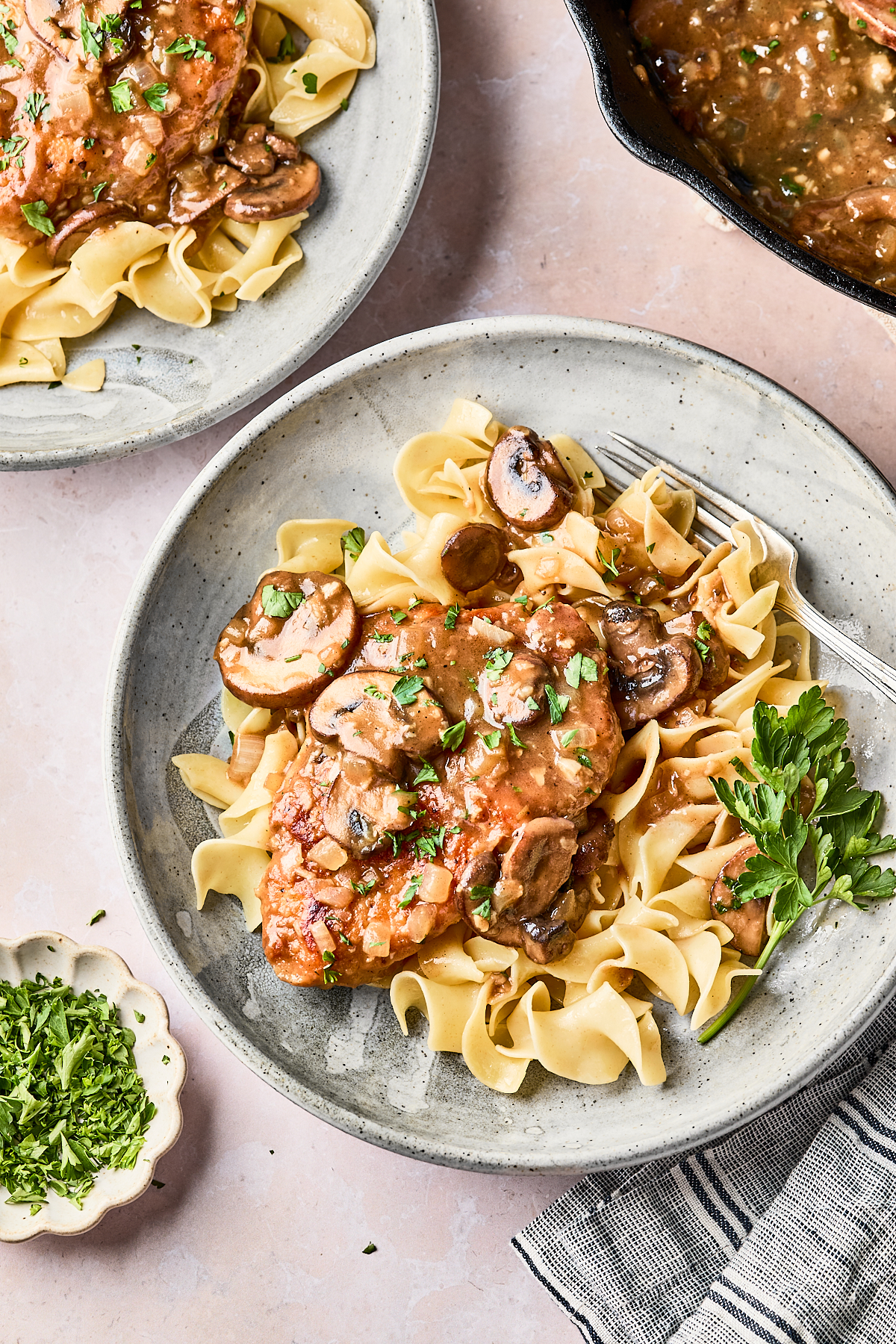 A plate of egg noodles topped with chicken and saut&eacute;ed mushrooms in a creamy sauce, garnished with chopped parsley. A fork rests on the plate, and fresh parsley and extra chopped herbs are nearby.