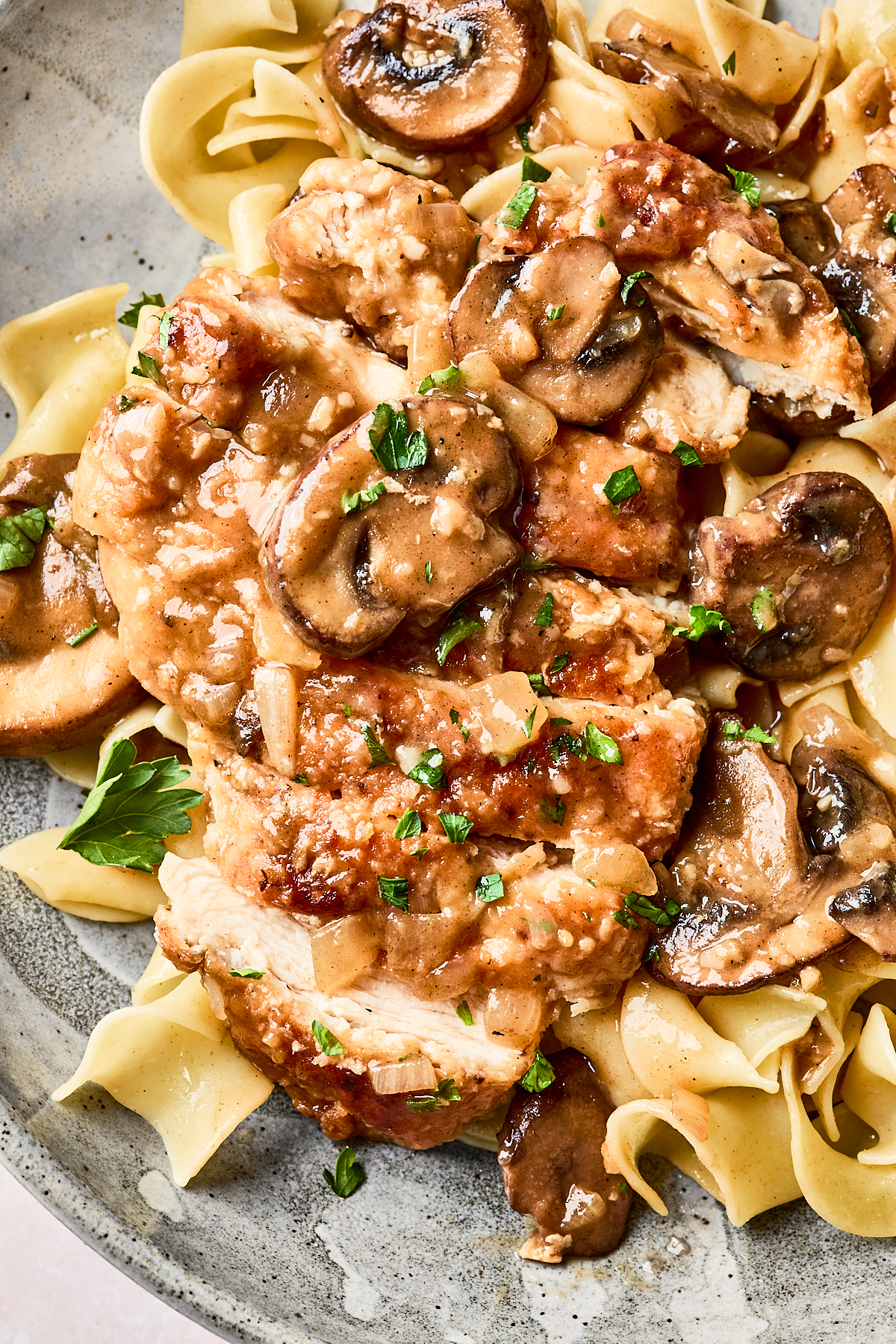A plate of egg noodles topped with sliced cooked chicken breast, saut&eacute;ed mushrooms, and a brown sauce, garnished with chopped parsley.