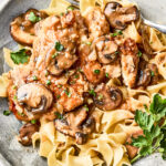 A plate of egg noodles topped with chicken and sautéed mushrooms in a creamy sauce, garnished with chopped parsley. A fork rests on the side of the plate.