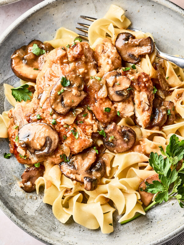 A plate of egg noodles topped with chicken and sautéed mushrooms in a creamy sauce, garnished with chopped parsley. A fork rests on the side of the plate.