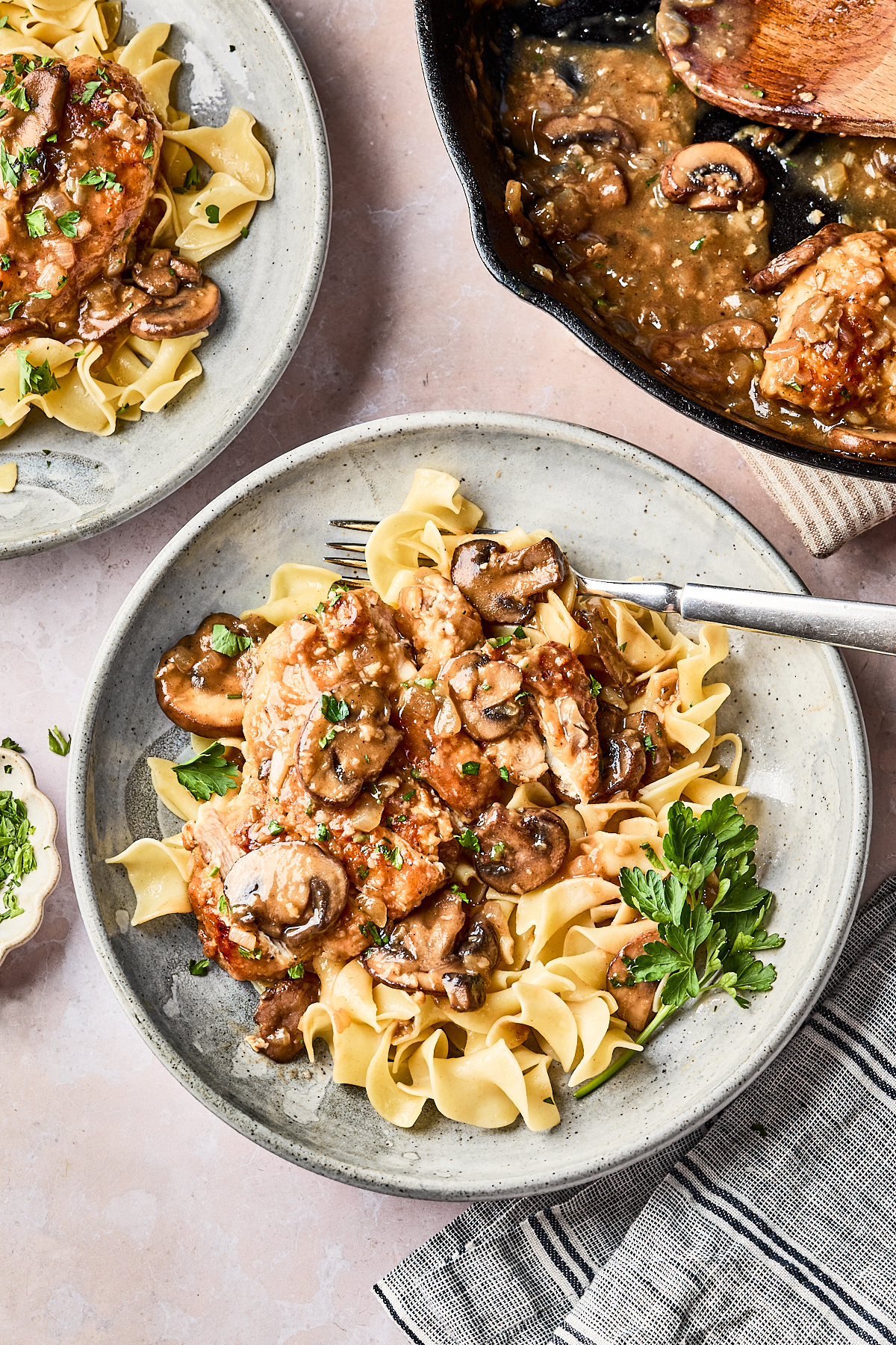 A plate of egg noodles topped with chicken, mushrooms, and a savory sauce, garnished with chopped parsley. A fork rests on the plate, and a skillet with more chicken and sauce is nearby.