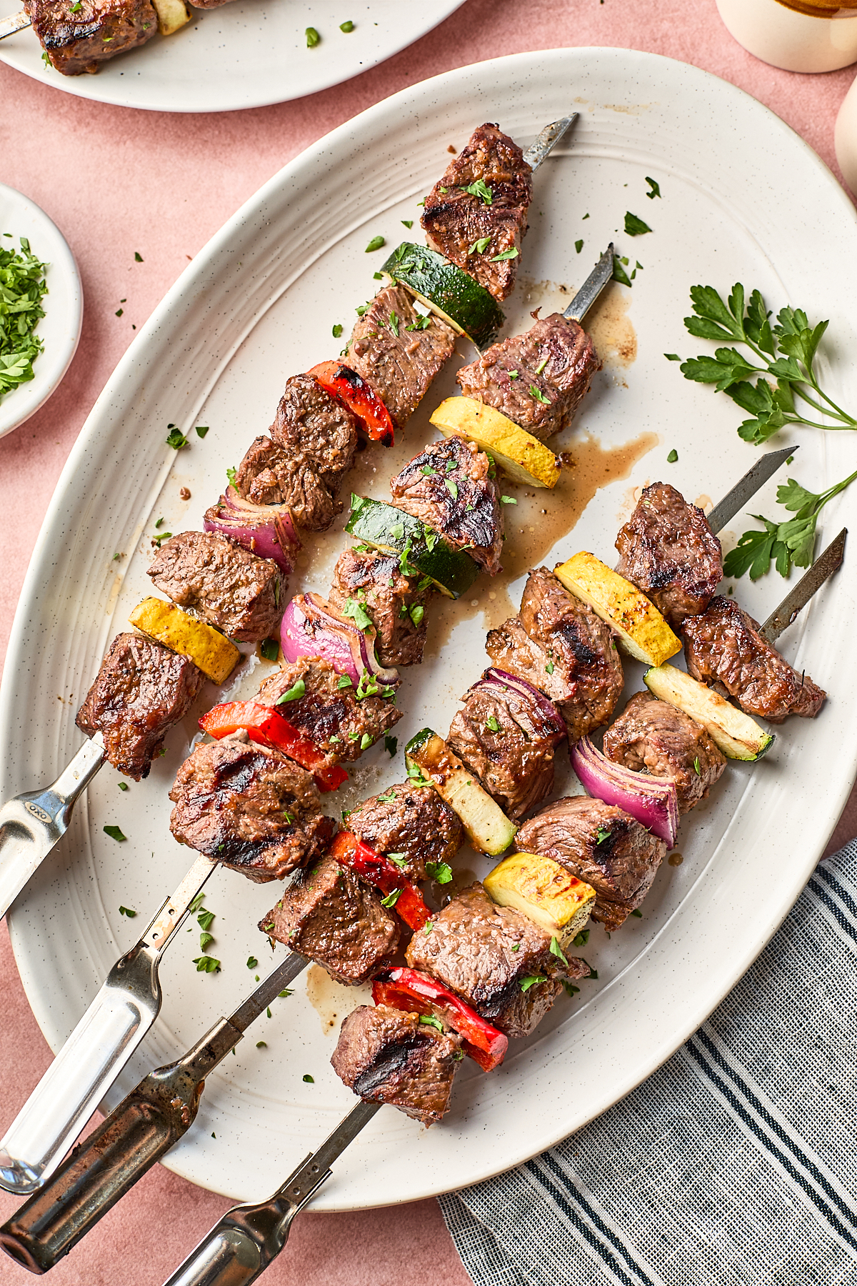 Three grilled beef skewers with colorful vegetables—red onion, red and yellow bell peppers, and zucchini—are served on a large white oval plate, garnished with chopped parsley. A striped napkin is visible at the side.
